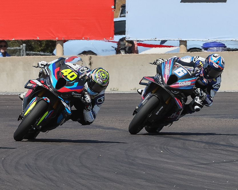 Two racers on motorcycles on a track with a red flag in the background